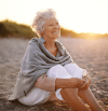 Eine ältere Frau sitzt lächelnd am Strand. 